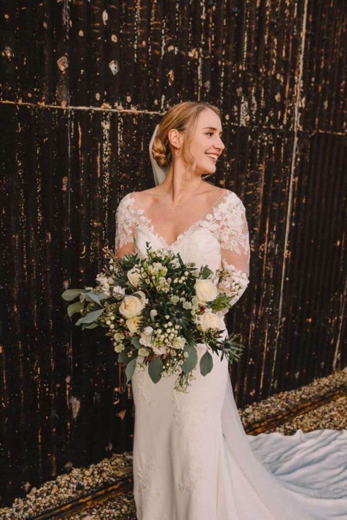 Bride smiling and looking away from the camera whilst holding flowers. Bridal hair and makeup by Bristol makeup artist Evie Smith