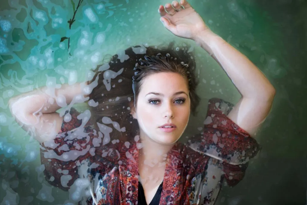 Model lies in a shallow pool of water with one arm above her head and her face just above the waterline to show off her perfect makeup.