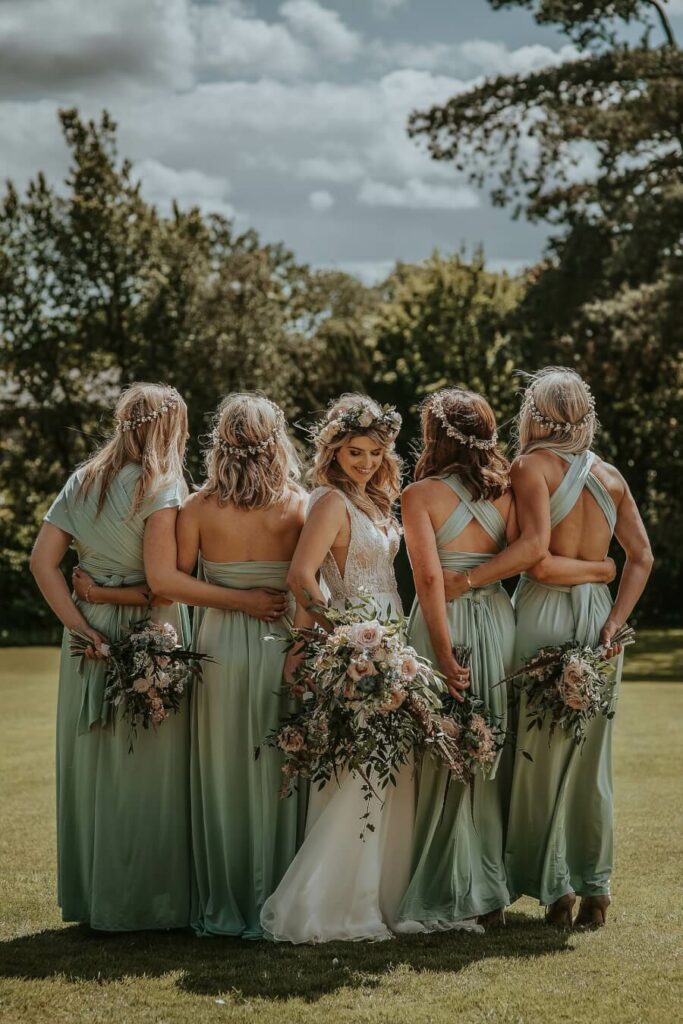 Bride standing with bridesmaids on a lawn, the bridesmaids are facing away from the camera as the bride looks over her shoulder.