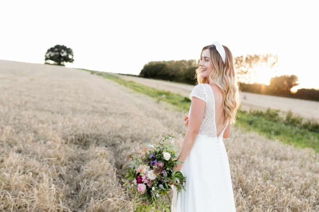 Bride in a field holding bouquet of flowers as the sun goes down in the background.