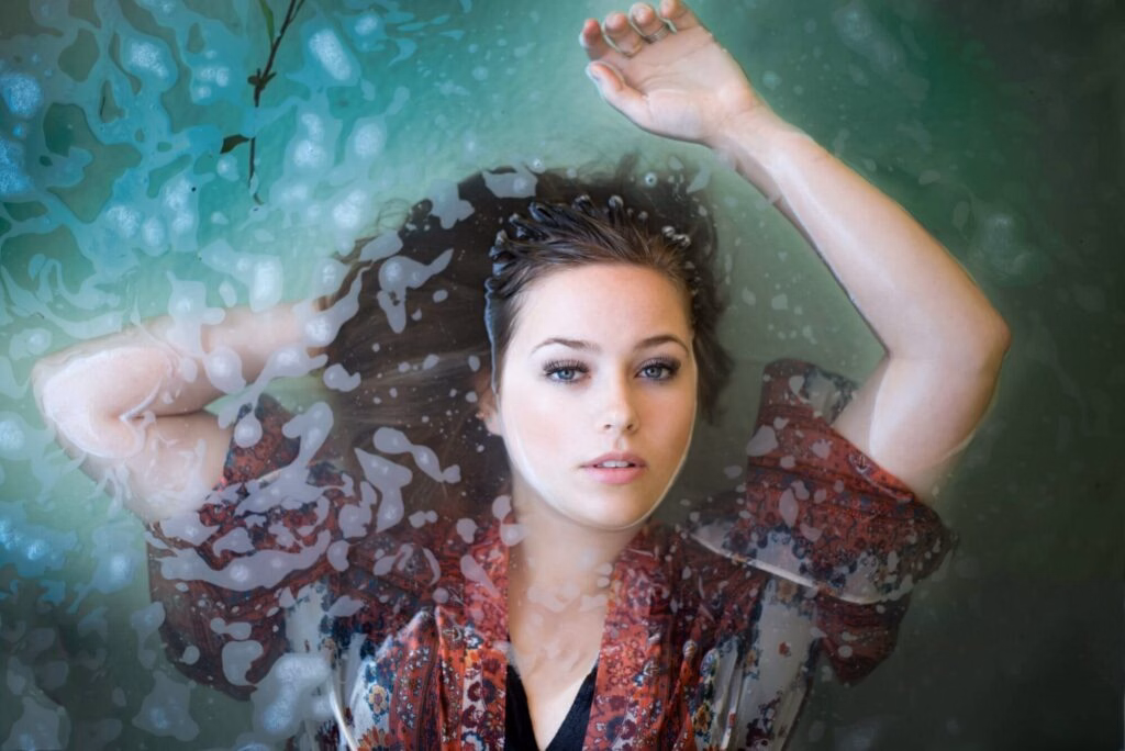 Model lies in a shallow pool of water with one arm above her head and her face just above the waterline to show off her perfect makeup.