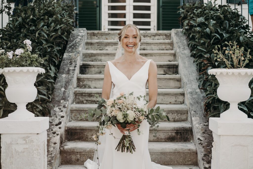 Bride dressed in white holding flowers standing at the bottom of the steps at a wedding venue in Italy. She is wearing white and smiling, staring straight into the camera.
