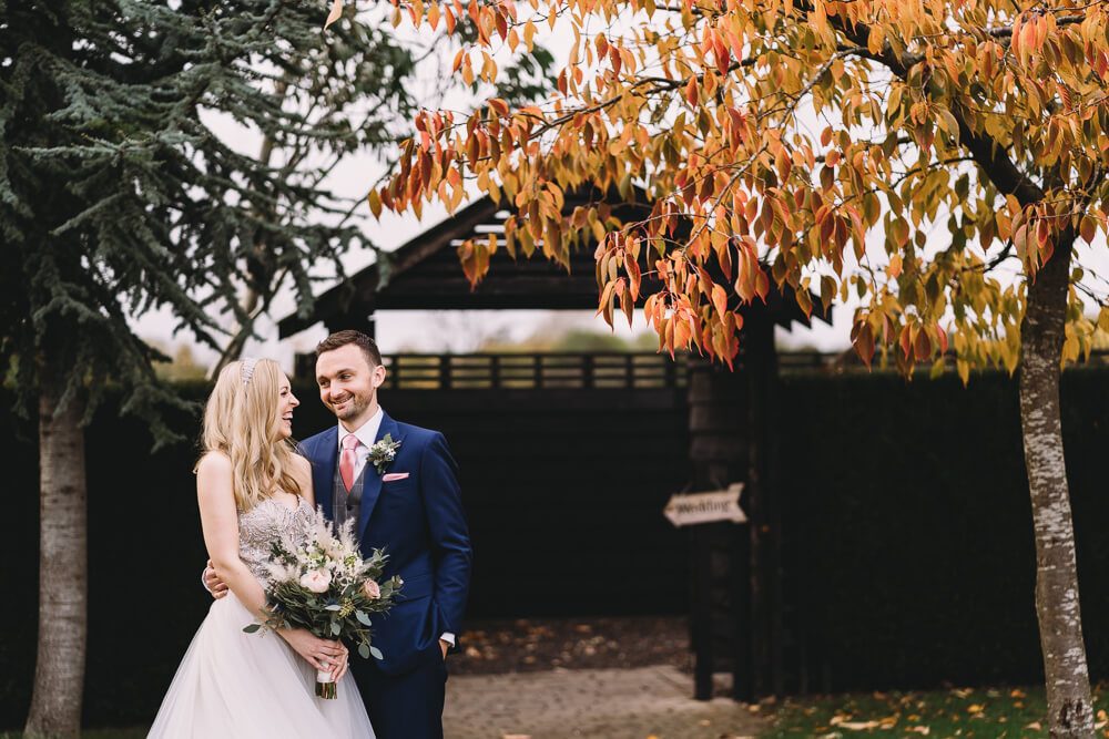 A smiling bride and groom outside by an autumn tree on their wedding day.