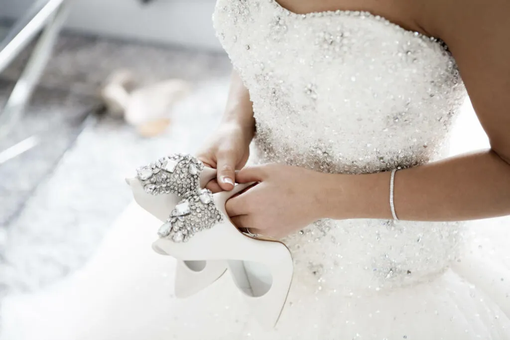 Stock photo of bride from the shoulders down wearing sparkly wedding dress and holding a pair of white sparkly high heels.