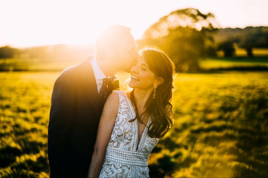 Groom kisses bride on the forehead on their wedding day as the sun goes down behind them. Bridal makeup by Bristol makeup artist Evie Smith.